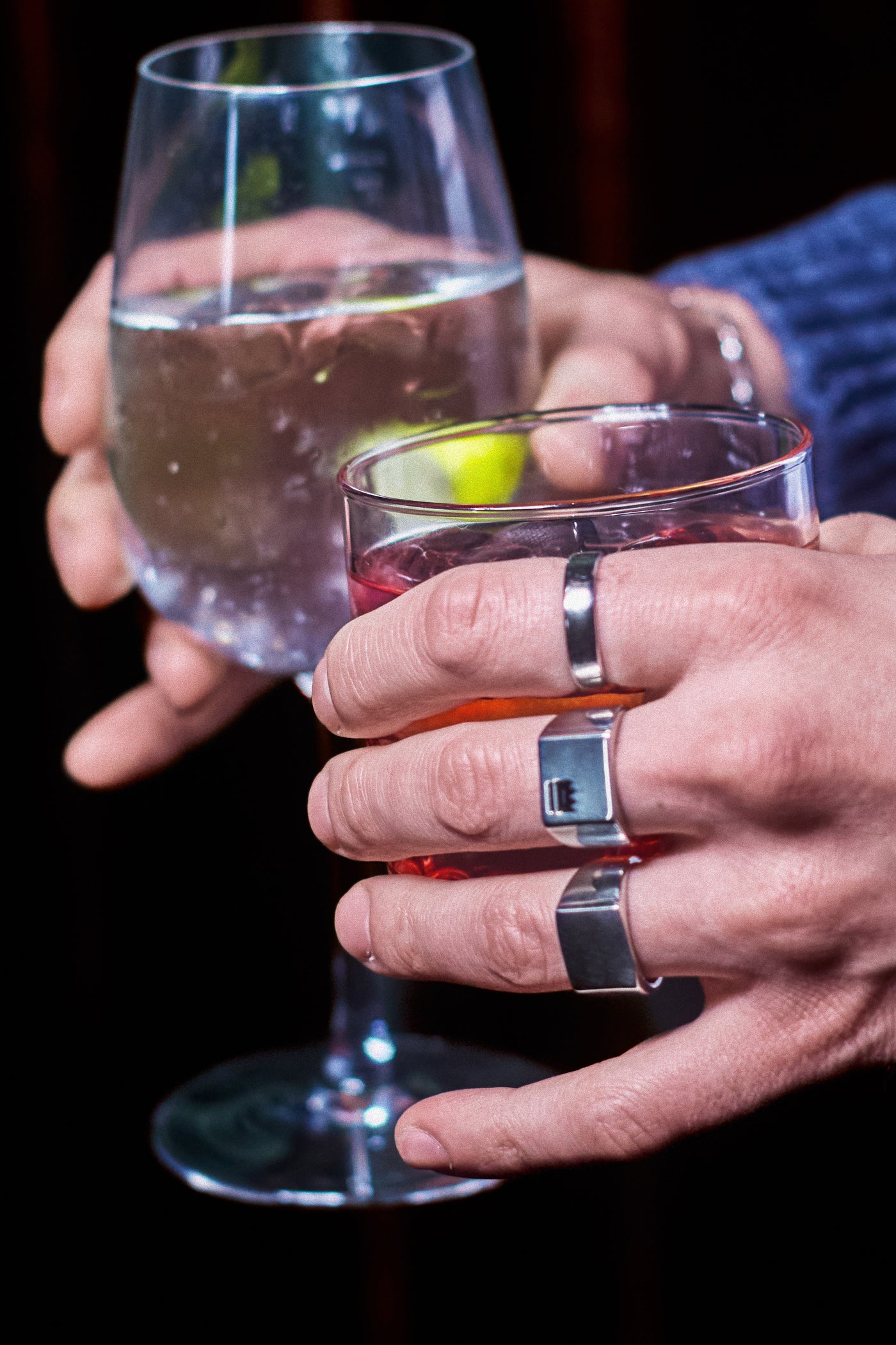 Plain Band Ring - stacked with Crown Signet Ring and Plain Signet Ring, two hands holding cocktail glasses, dark editorial setting