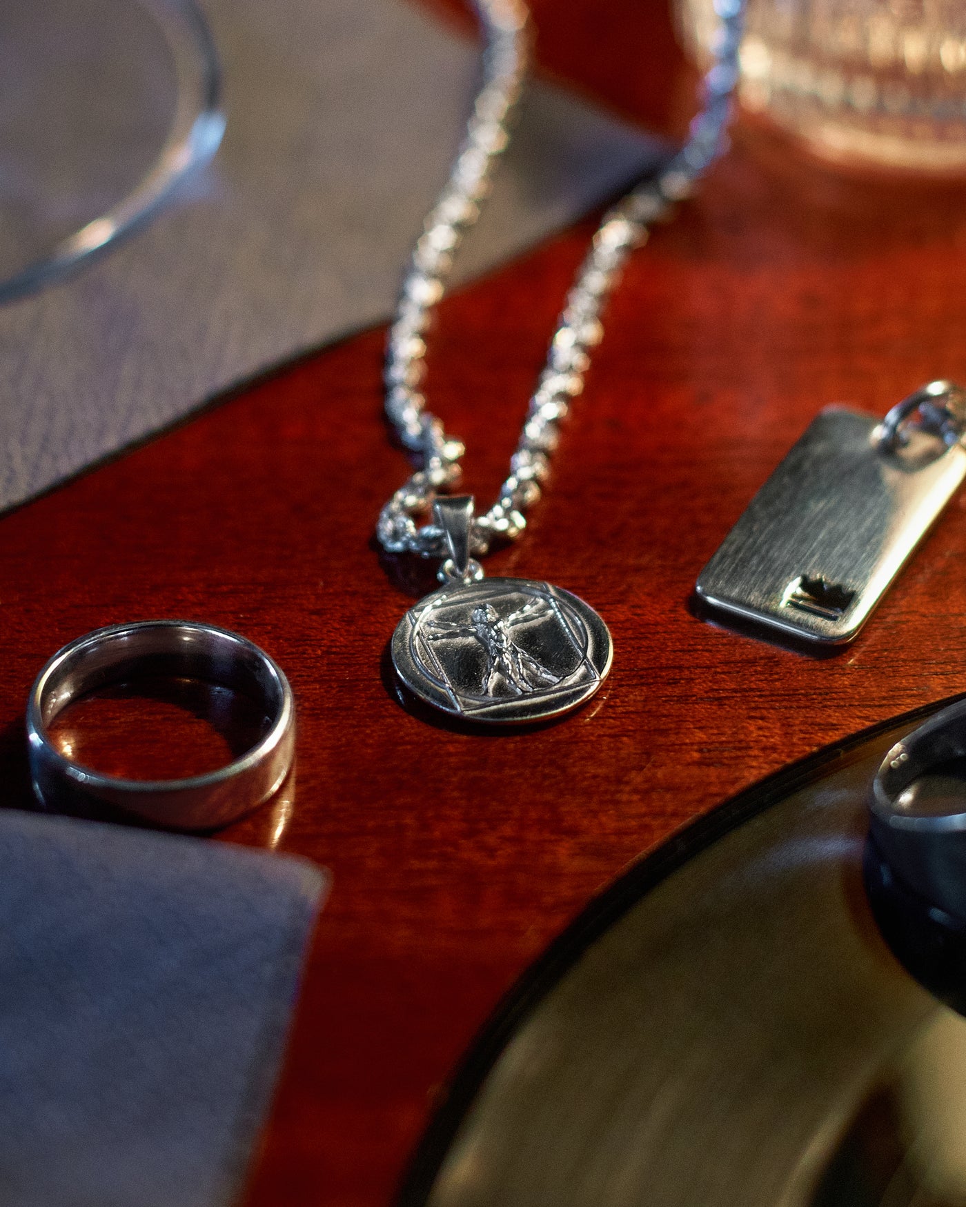 Vitruvian Pendant - styled with Plain Band Ring and Plain Dog Tag, tabletop arrangement on dark wood with cocktail glasses framing