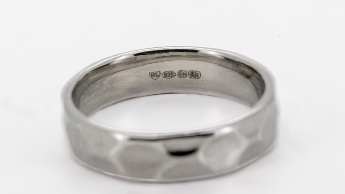 A silver-toned hammered band ring with a polished inner band showing sterling silver purity stamps, set against a plain white background.